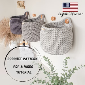 Può includere: Tre cesti appesi all'uncinetto in grigio e viola con manici in legno. I cesti sono appesi a una parete bianca con ganci in legno. Il testo "CROCHET PATTERN PDF & VIDEO TUTORIAL" è in un cerchio nero su sfondo bianco.