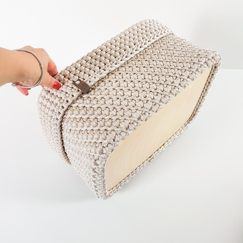 May include: A beige crocheted storage basket with a wooden base. The basket has a decorative brown leather strap.