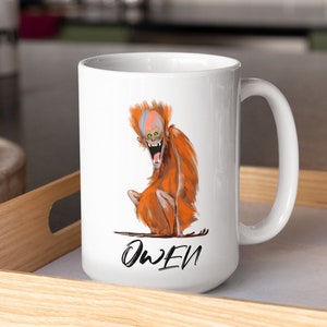 May include: White ceramic coffee mug with a cartoon illustration of an orange-haired creature with a wide open mouth. The creature is sitting on a branch. The mug has the word "Owen" written in black at the bottom.