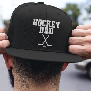 May include: Black baseball cap with a white embroidered design that says "Hockey Dad" with two crossed hockey sticks.