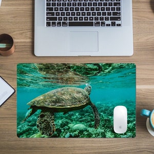 May include: A green and blue mouse pad with a picture of a sea turtle swimming underwater. The turtle is brown and green with a light blue background. The mouse pad is on a wooden desk with a laptop, a notebook, a cup of coffee, and a book.