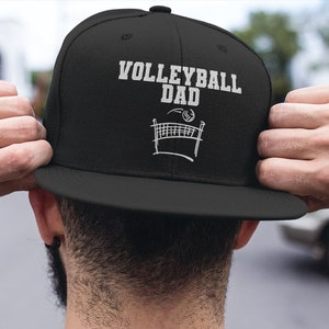 May include: Black baseball cap with white text that reads "Volleyball Dad" and a graphic of a volleyball net with a volleyball on top.