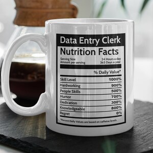 Może przedstawiać: Biały ceramiczny kubek z czarno-białym nadrukiem etykiety wartości odżywczych dla "Data Entry Clerk". Etykieta zawiera listę różnych umiejętności i ich procentową dzienną wartość, np. "Poziom umiejętności" na 1000% i "Żal" na 0%.