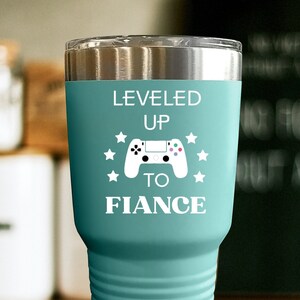 May include: A teal blue tumbler with a stainless steel lid and a white design that says "LEVELED UP TO FIANCE" with a video game controller graphic.