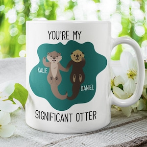 Könnte beinhalten: Eine weiße Keramiktasse mit einem blaugrünen Hintergrund, auf dem zwei Cartoon-Otter Hand in Hand halten. Der Text "You're my significant otter" ist auf die Tasse gedruckt. Die Otter sind mit "Kalie" und "Daniel" beschriftet.