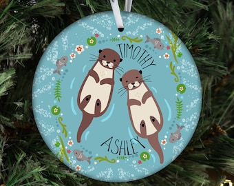 May include: A blue and white ceramic Christmas ornament featuring two cartoon otters swimming in a blue pond. The ornament has the names "Timothy" and "Ashley" written on it.
