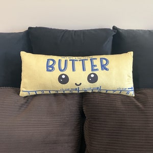 May include: A yellow rectangular cushion with the word "BUTTER" in blue, featuring a smiling face design. The cushion has a measuring guide along the bottom and is labeled "NET WT. 4 OZ (113g)".