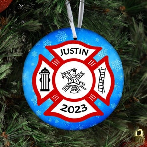 May include: A blue and red Christmas ornament with a firefighter's emblem, a fire hydrant, a ladder, and a helmet. The ornament has a white snowflake pattern and the text "JUSTIN 2023".