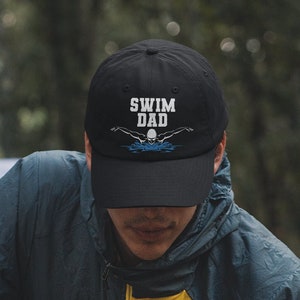May include: A person wearing a black baseball cap with the text "Swim Dad" and a swimmer graphic in white. The person is sitting by a campfire, holding a lighter and starting a fire.