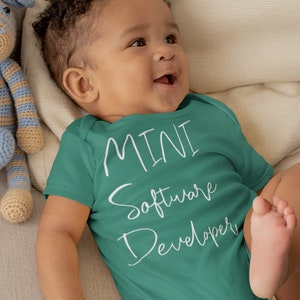May include: A baby wearing a teal bodysuit with the text "MINI Software Developer" in white. The baby is lying on a light brown couch with two crocheted stuffed animals.