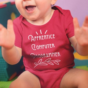 May include: A baby wearing a red onesie with the text "I/ Apprentice Computer Programmer" in white. The baby is sitting on a colorful play mat and smiling.