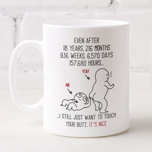 May include: White ceramic mug with a humorous graphic and text. The mug features a cartoon illustration of two figures and the phrase "EVEN AFTER 18 YEARS... I STILL JUST WANT TO TOUCH YOUR BUTT. IT'S NICE." A white vase with yellow flowers is next to the mug.
