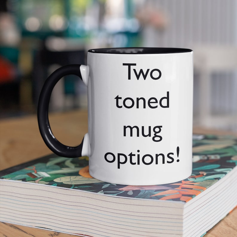 K&ouml;nnte beinhalten: Eine wei&szlig;e Keramiktasse mit schwarzem Henkel und schwarzem Rand. Auf der Tasse steht der Text "Two toned mug options!" gedruckt.