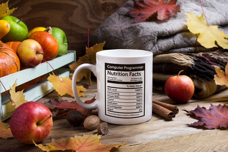 K&ouml;nnte beinhalten: Eine wei&szlig;e Kaffeetasse mit einer schwarz-wei&szlig;en Grafik, auf der "Computer Programmer Nutrition Facts" steht, mit einer Liste von F&auml;higkeiten und deren prozentualem Tageswert. Die Tasse steht auf einem Holztisch mit Herbstbl&auml;ttern, &Auml;pfeln und Zimtstangen.