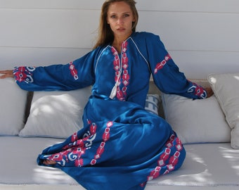 Könnte beinhalten: Eine Frau trägt ein langes, blaues Kleid mit weißen und rosa Stickereien. Sie sitzt auf einem weißen Sofa mit weißen Kissen.