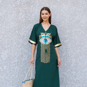 May include: Emerald green maxi dress with a V-neck and short sleeves. The dress features an eye-shaped design with gold and blue embellishments on the front. Gold trim accents the sleeves. A straw tote bag with a white rope handle is held in the hand.