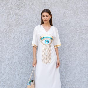 May include: White maxi dress with a V-neck and short sleeves. The dress features a large eye design in blue and gold sequins on the front. Gold trim accents the sleeves. A straw tote bag and black heels complete the look.