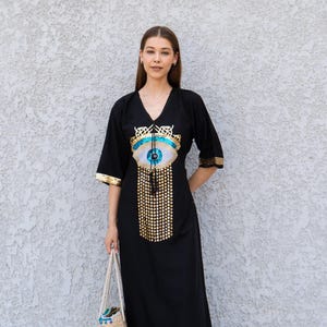 Peut inclure: Longue robe noire avec un col en V et des manches aux coudes. La robe présente un grand motif d'œil avec des ornements dorés et bleus. Le modèle tient un sac fourre-tout tissé.