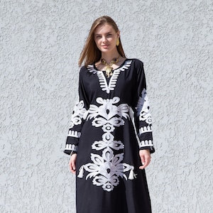 Pode incluir: Um vestido maxi preto com desenhos florais bordados em branco e um detalhe de borla. O vestido tem um decote em V e mangas compridas.