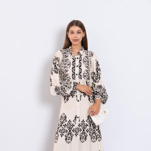 Pode incluir: Um vestido longo creme com um padrão floral preto. O vestido apresenta uma frente abotoada, mangas compridas e uma saia fluida. Uma bolsa branca com uma alça de corrente é segurada pela pessoa que usa o vestido.