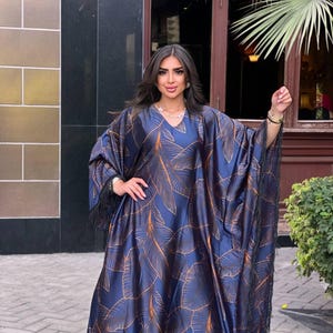May include: A flowing, navy blue caftan dress with a gold leaf pattern. The dress features a V-neck and fringe trim along the sleeves and hem. The fabric has a silky sheen, and the overall design is elegant and sophisticated.