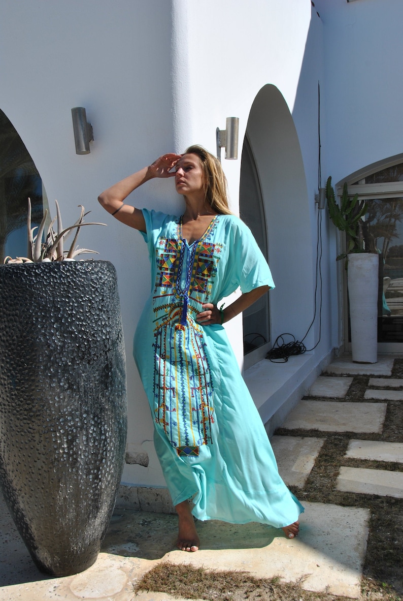May include: A woman wearing a light blue maxi dress with colorful embroidery. The dress has a V-neckline and short sleeves. She is standing in front of a white wall with an arched doorway.
