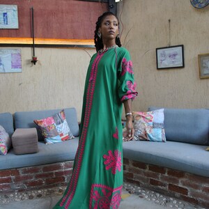 Puede incluir: Una mujer lleva un vestido maxi verde con bordados rosas. El vestido tiene una falda larga y fluida y un corpi&ntilde;o ajustado. La mujer est&aacute; de pie en un patio con una pared de ladrillo y un sof&aacute; en el fondo.