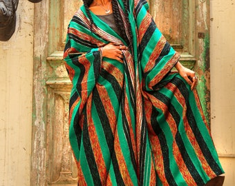 May include: A woman wearing a long, green, black, and orange striped robe with a silver necklace. The robe is draped over one shoulder and the woman is standing in front of a wooden door.