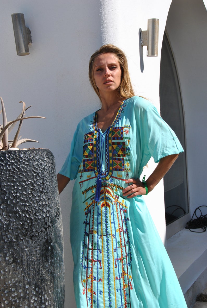 May include: A light blue, long-sleeved dress with colorful embroidery. The dress has a V-neck and a tassel hanging from the neckline.