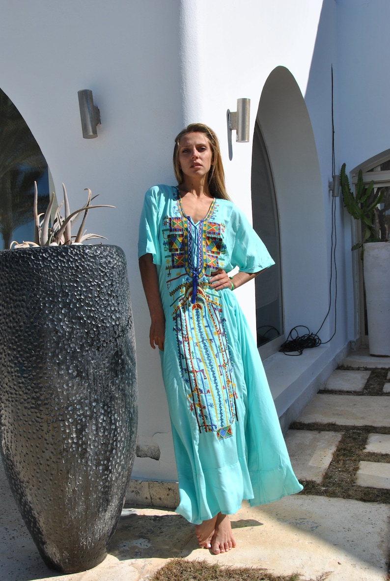 May include: A woman wearing a light blue maxi dress with colorful embroidery. The dress has a V-neckline and short sleeves. She is standing in front of a white wall with a potted plant.