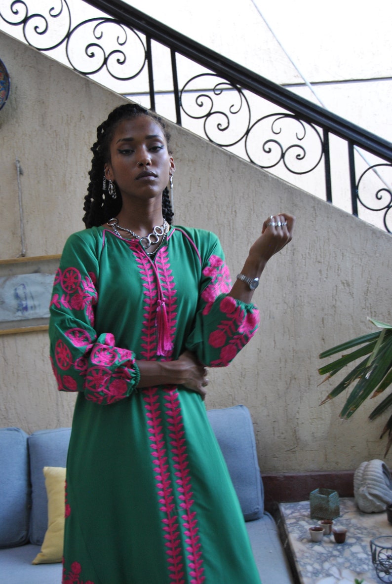 Puede incluir: Una mujer lleva un vestido verde con bordados rosas. El vestido tiene mangas largas y un borla en el escote. Lleva joyas de plata y un reloj.