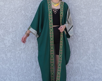May include: A long, green kimono with gold and green embroidery. The kimono has a black and white geometric pattern on the sleeves. The model is wearing a black top and black shorts.