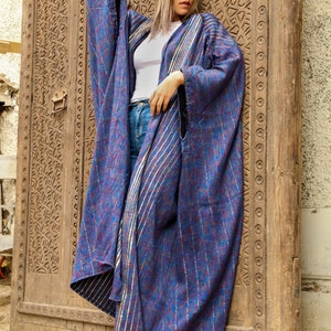 Op de afbeelding: Een vrouw draagt een lange, blauw-paarse gestreepte robe met gouden accenten. De robe is losjes gesneden en wordt gedragen over een wit shirt en een blauwe jeans. De vrouw staat voor een houten deur met ingewikkelde houtsnijwerk.