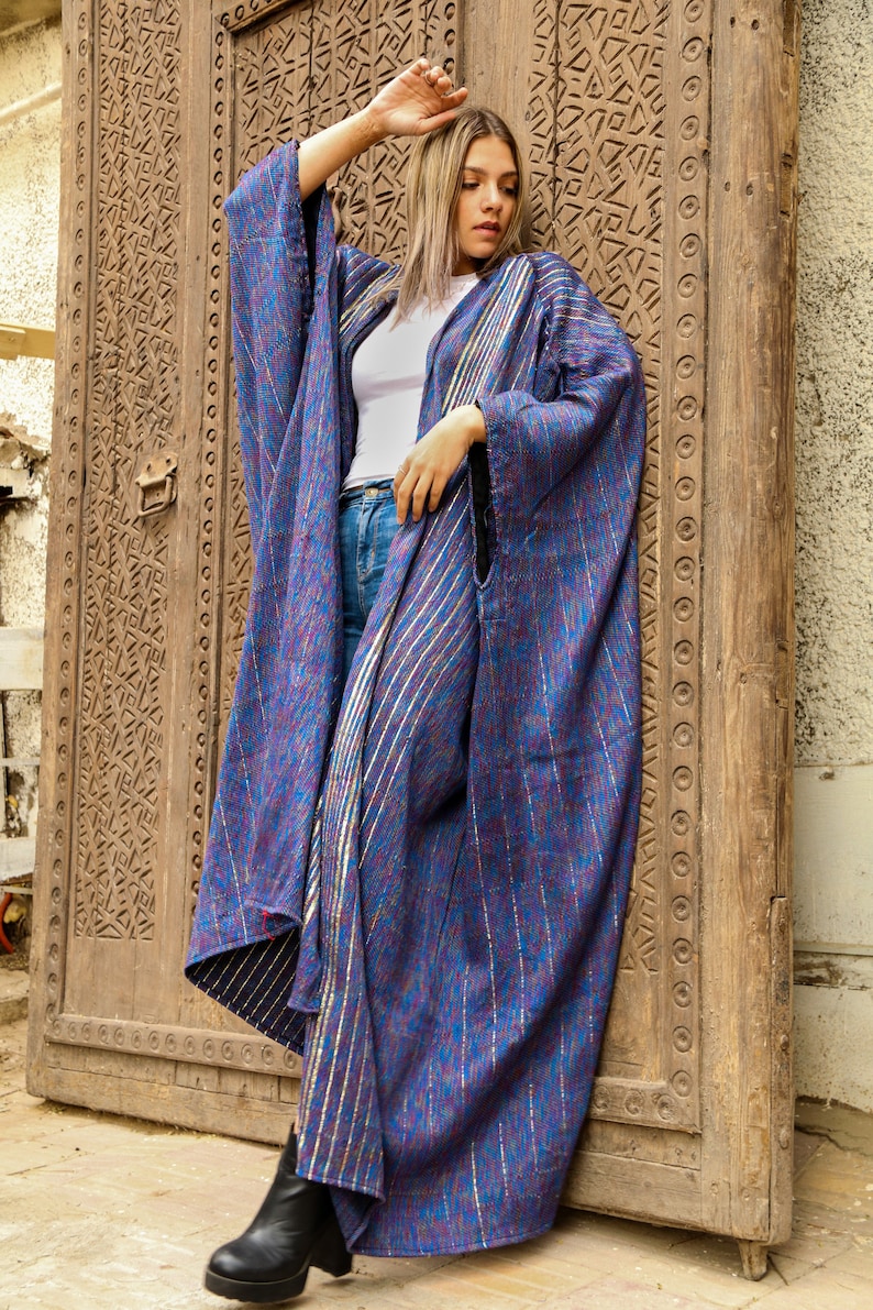 Op de afbeelding: Een vrouw draagt een lange, blauw-paarse gestreepte robe met gouden accenten. De robe is losjes gesneden en wordt gedragen over een wit shirt en een blauwe jeans. De vrouw staat voor een houten deur met ingewikkelde houtsnijwerk.