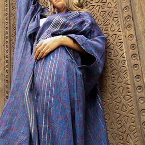 Op de afbeelding: Een vrouw draagt een lange, blauw-paarse gestreepte robe met zilveren accenten. De robe is over haar lichaam gedrapeerd en ze staat voor een houten deur met ingewikkelde houtsnijwerk.