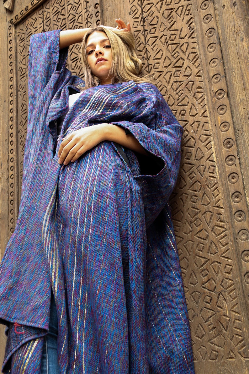 Op de afbeelding: Een vrouw draagt een lange, blauw-paarse gestreepte robe met zilveren accenten. De robe is over haar lichaam gedrapeerd en ze staat voor een houten deur met ingewikkelde houtsnijwerk.