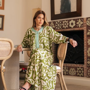 May include: A woman wearing a green and white patterned dress with a light blue trim. She is sitting on a chair in a room with a fireplace and a painting on the wall.