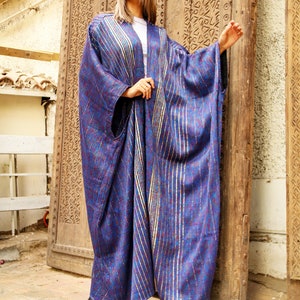 Op de afbeelding: Een lange, vloeiende blauwe en paarse robe met gouden accenten. De robe heeft een diagonaal streepdessin en wordt gedragen door een persoon die voor een houten deur staat.
