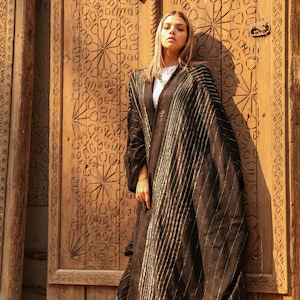 Puede incluir: Una mujer con una abaya negra con bordados plateados se encuentra frente a una puerta de madera tallada. La abaya tiene una silueta larga y fluida y está adornada con detalles intrincados. La puerta está tallada con intrincados patrones geométricos.