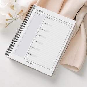 May include: A white spiral-bound planner with a "WEEKLY MENU" header and sections for each day of the week. It includes a grocery list section. The planner is open, revealing blank pages for notes and planning.