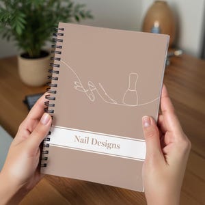 May include: A spiral-bound notebook with a taupe cover featuring a white line drawing of a hand and nail polish bottle. The words "Nail Designs" are printed in a white banner across the bottom. The notebook is held by a person.