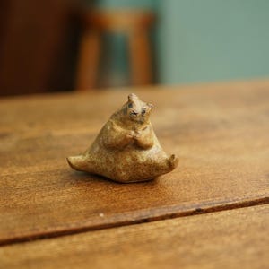 May include: A small, tan ceramic figurine of a seated animal with its paws clasped. The figurine has a rounded body and a small head with visible eyes. It is placed on a wooden surface.