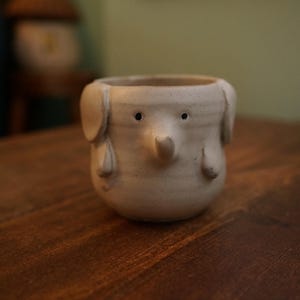 May include: A small, off-white ceramic planter shaped like an elephant. The planter has a round body, large ears, a trunk, and small black eyes. The planter is sitting on a wooden surface.