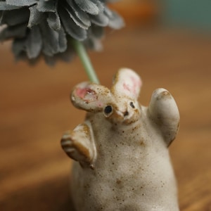 Può includere: Una stravagante statuetta di coniglio in ceramica con orecchie foderate di rosa e una finitura bianco sporco macchiettata. Il coniglio tiene uno stelo che sostiene un fiore di stoffa grigia. È un oggetto decorativo.
