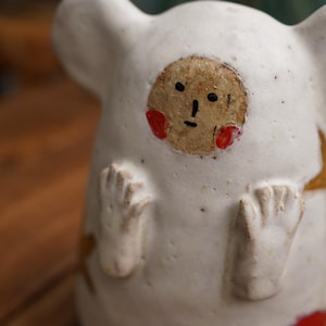 Puede incluir: Figura de cerámica hecha a mano con forma de criatura con cuerpo blanco y cara marrón con mejillas rojas. La figura tiene brazos y pies pequeños, y un diseño de estrella dorada en el lateral.
