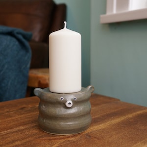 Könnte beinhalten: Eine weiße Säulenkerze steht auf einem handgefertigten, grauen Kerzenhalter aus Keramik. Der Kerzenhalter hat ein bärenähnliches Design mit kleinen, runden Augen und einer Schnauze. Die Kerze und der Halter stehen auf einer Holzoberfläche.