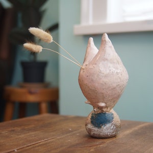 Puede incluir: Un jarrón de cerámica hecho a mano con una combinación de colores rosa claro y azul. El jarrón tiene una forma única y abstracta con dos formas que apuntan hacia arriba. Contiene tres tallos beige secos y esponjosos. El jarrón está sobre una superficie de madera.