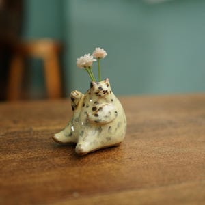 Könnte beinhalten: Eine katzenförmige Keramikvase mit gesprenkelter, hellgrüner Glasur. Die Vase enthält zwei kleine, weiße Blumen. Die Katze sitzt mit ihren Pfoten nach vorne auf einer Holzoberfläche.