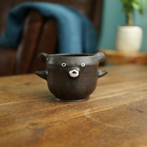 May include: A dark brown ceramic mug shaped like a bear with two small ears and two side arms extending from the body. The mug features a painted white face with two circular eyes and a small nose area centered on the front of the rounded vessel.