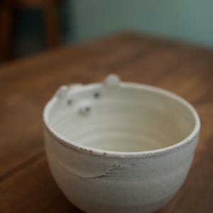 Könnte beinhalten: Eine handgefertigte, cremefarbene Keramikschale mit einem bärenartigen Design am Rand. Die Schale hat eine strukturierte Oberfläche und eine leicht unregelmäßige Form, was auf einen handwerklichen Ursprung hindeutet. Die Schale steht auf einer Holzoberfläche.